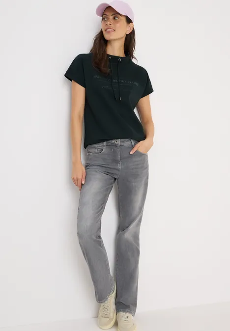 Frau mit schwarzem Shirt, grauer Jeans und rosa Kappe lehnt an der Wand.